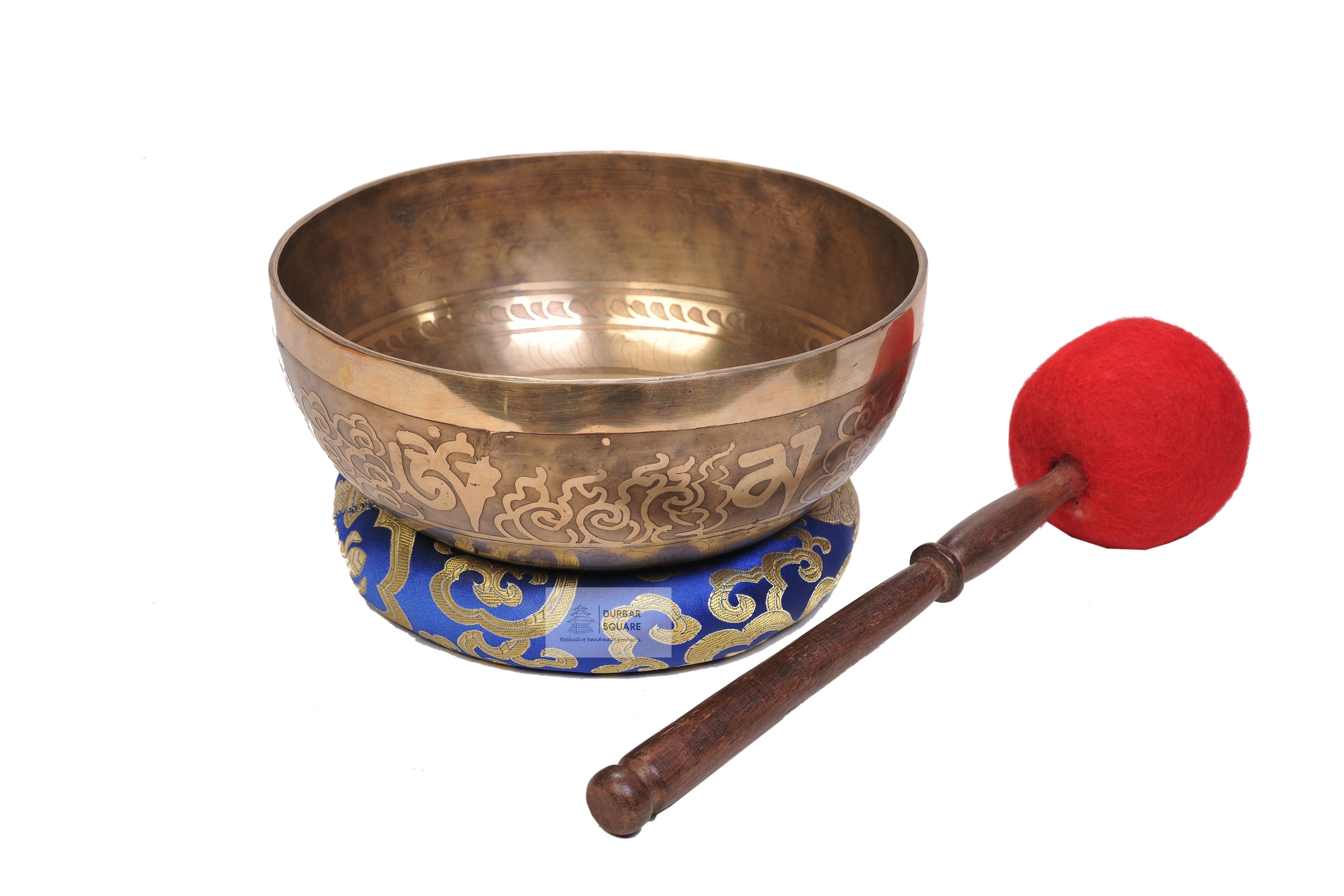 Polished gold-toned singing bowl engraved with the Om Mani Padme Hum mantra, on a blue brocade cushion with a red felt-tipped striker