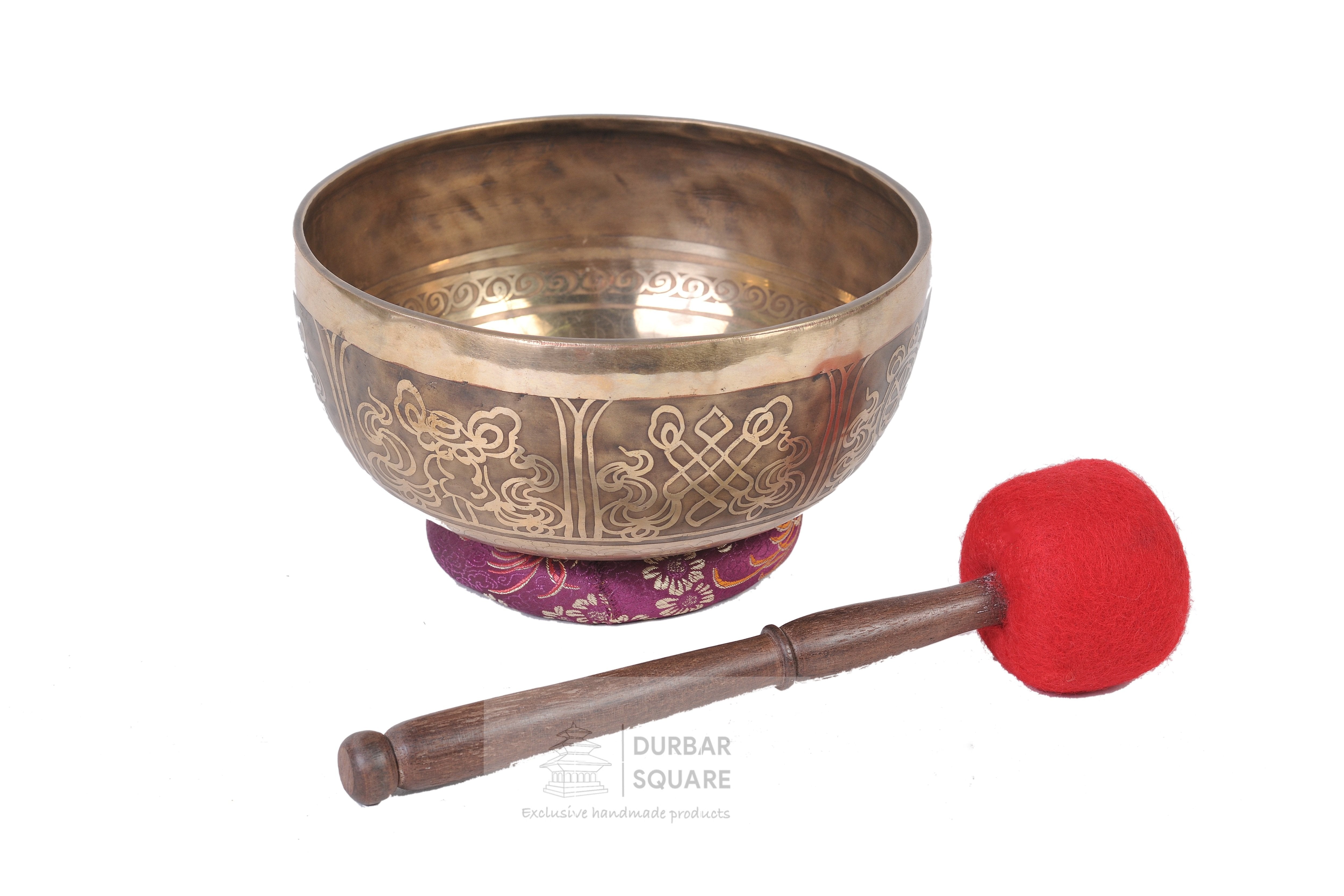 Brass singing bowl engraved with endless knot motifs, resting on a brocade cushion with a mallet beside it